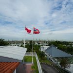 Two flagpoles flying Singapore and Norway flags beside white fence near rooftops and trees under cloudy sky