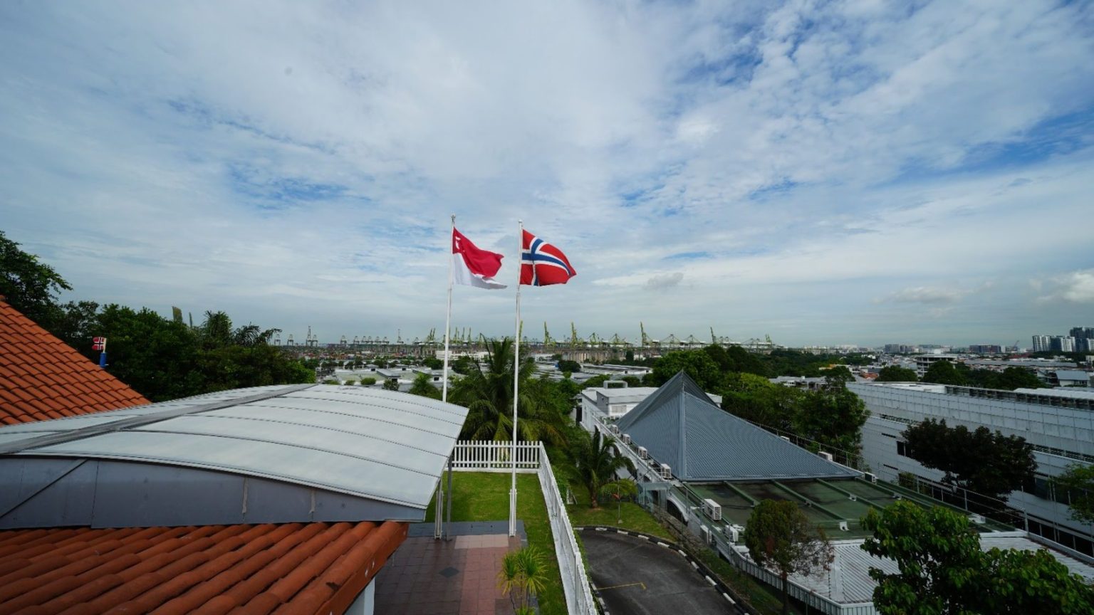 Two flagpoles flying Singapore and Norway flags beside white fence near rooftops and trees under cloudy sky