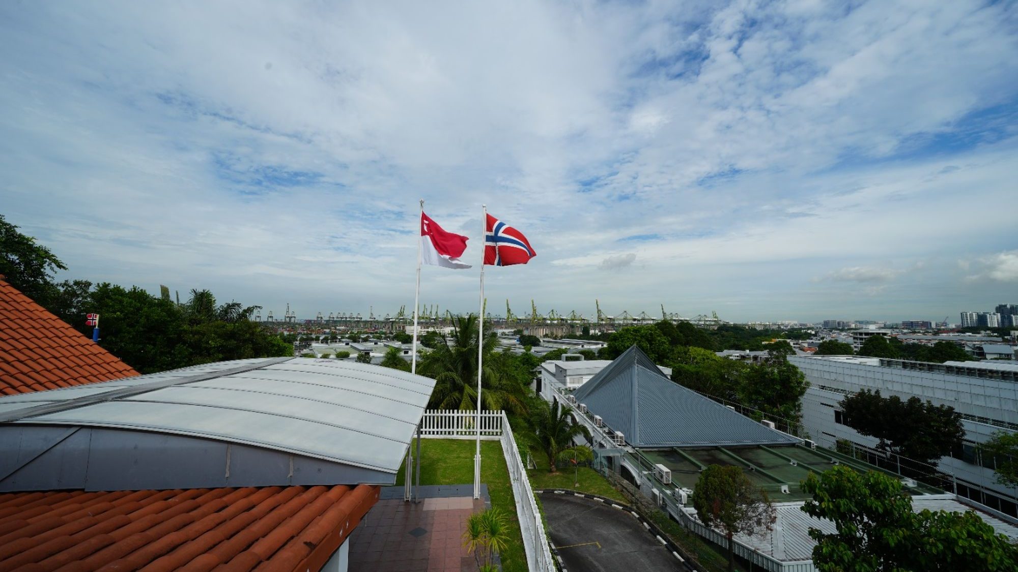 Two flagpoles flying Singapore and Norway flags beside white fence near rooftops and trees under cloudy sky