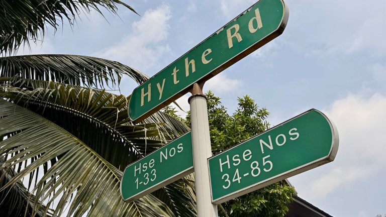 Green street signs for Hythe Road and house numbers 1-33 and 34-85 against a blue sky and palm leaves