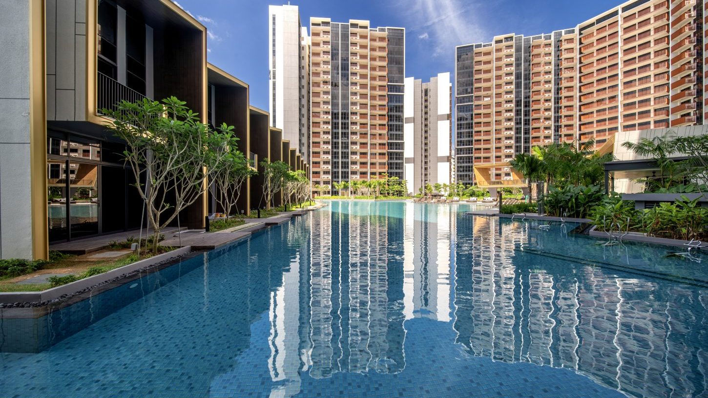 Residential towers reflected in a large swimming pool with greenery and poolside seating under blue sky