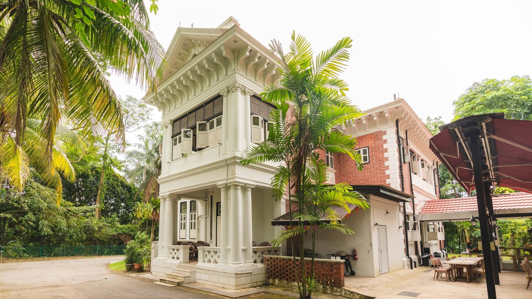 Two-storey colonial style house with white pillars and red brick walls surrounded by tall trees and greenery