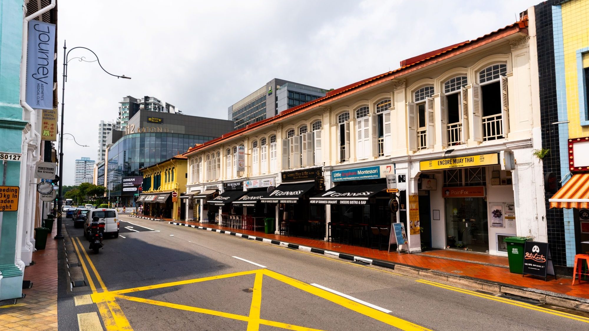 Shophouses with restaurants and clinics along a street with vehicles and modern buildings in the background