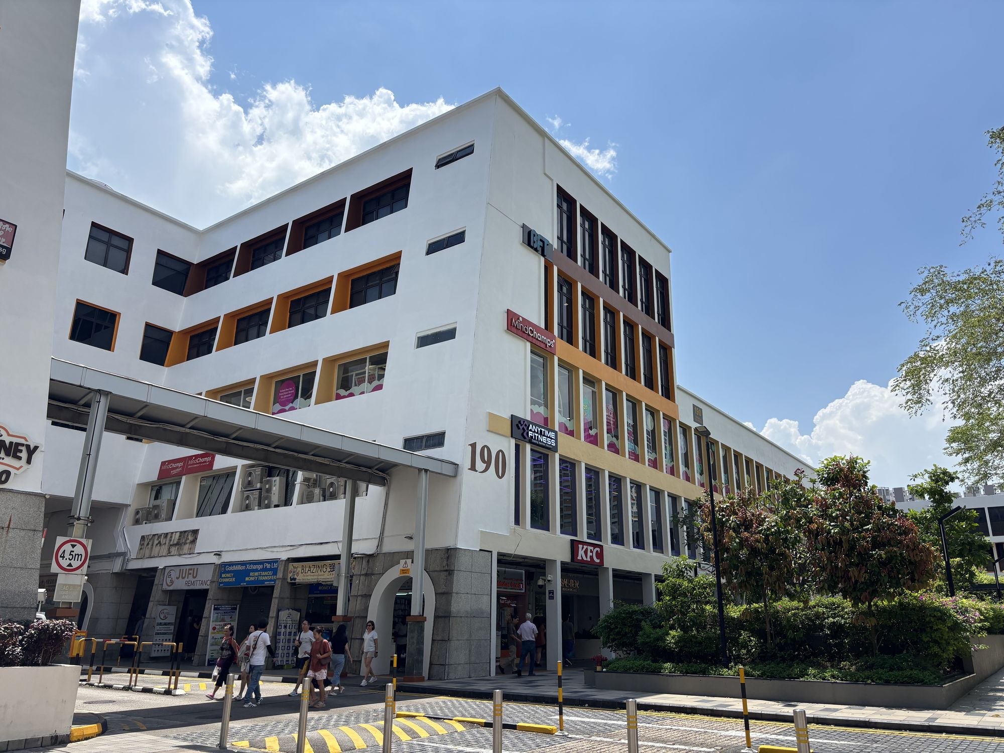 Mid-rise commercial building with shops including KFC, Anytime Fitness, and remittance services under clear blue sky
