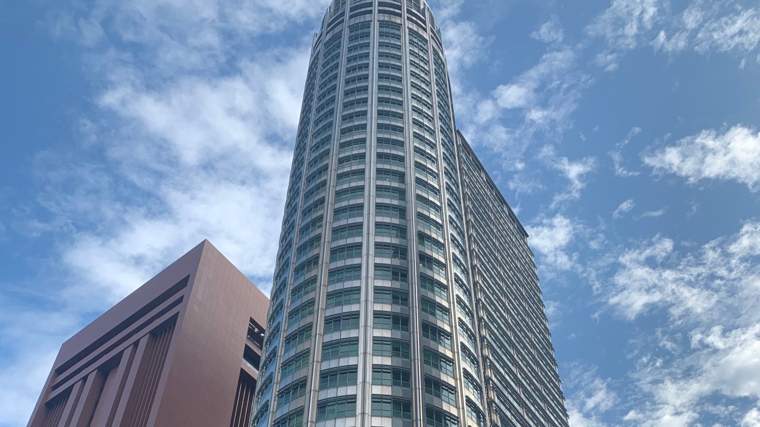 Tall modern office tower with glass windows next to a brown block under a blue sky with clouds