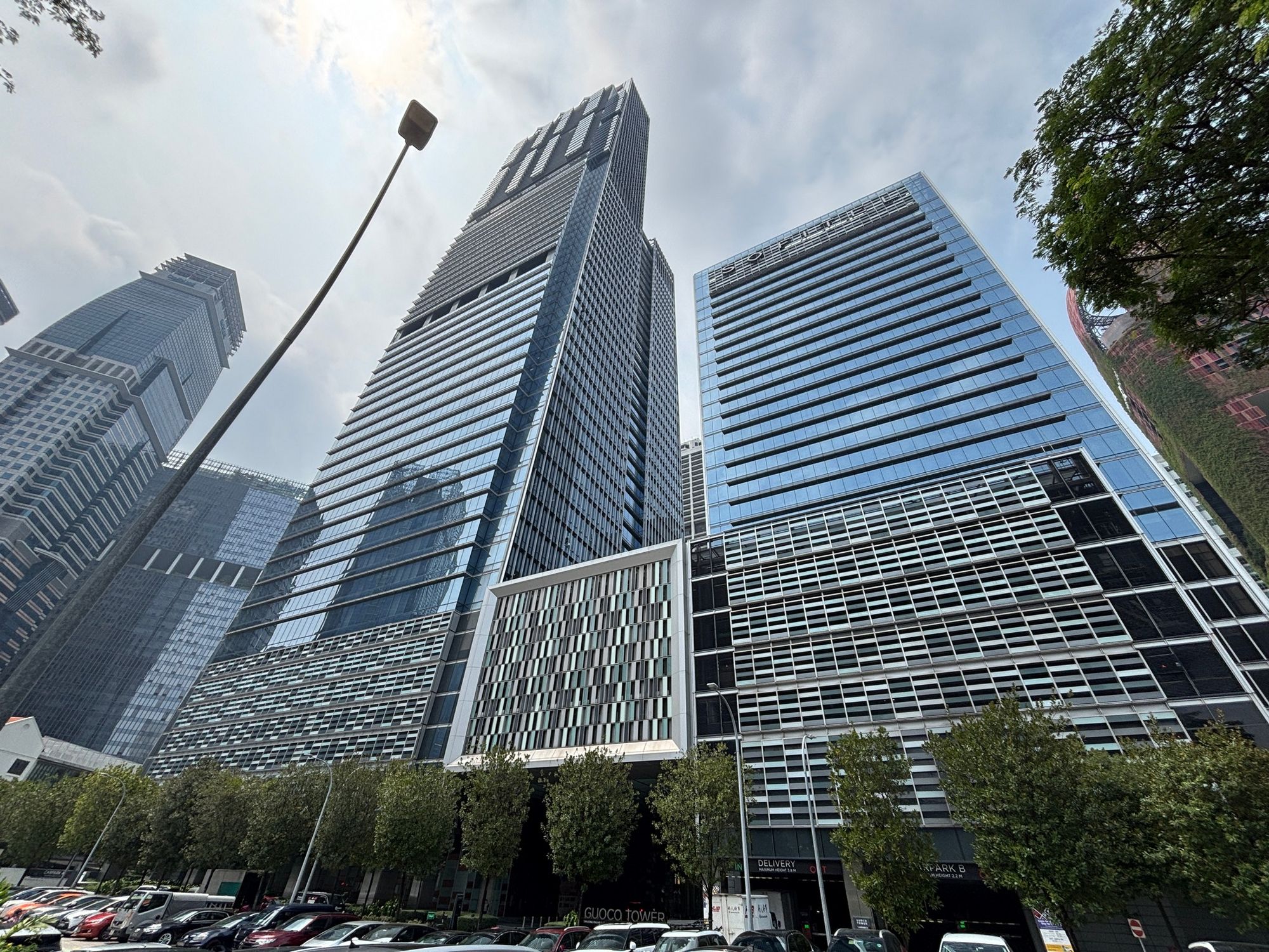 Tall glass office towers with parking lot and leafy trees at the base against cloudy sky