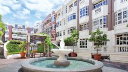 Fountain in front of low-rise residential blocks with white windows and balcony railings under blue sky