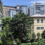 Residential blocks with balconies and greenery beside a large cream-coloured building under blue sky
