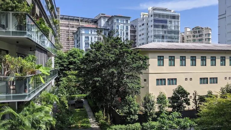 Residential blocks with balconies and greenery beside a large cream-coloured building under blue sky