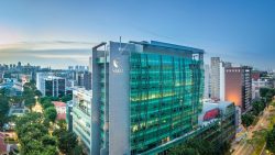 Tall modern building with green glass facade and SMU logo, surrounded by city skyscrapers and trees at dusk