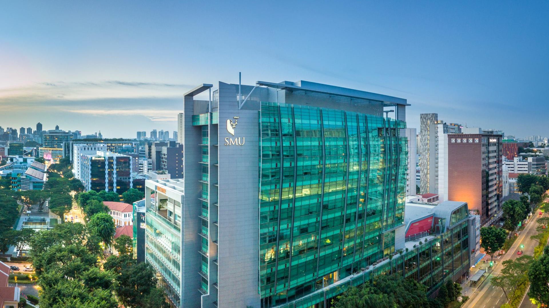 Tall modern building with green glass facade and SMU logo, surrounded by city skyscrapers and trees at dusk