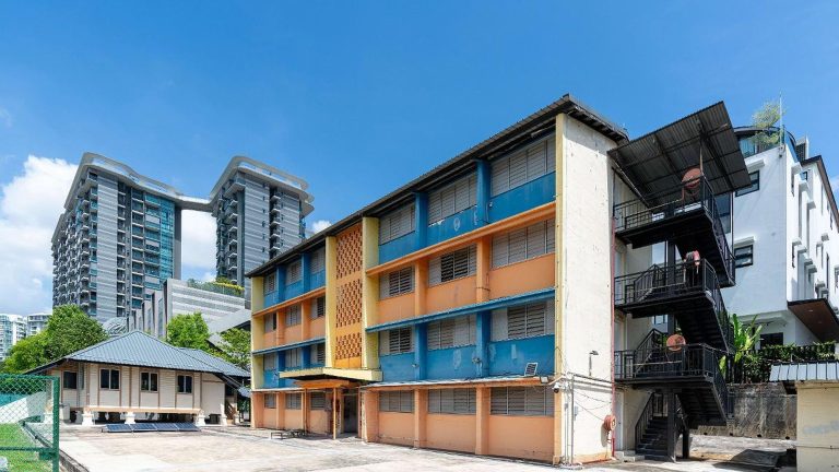 Three-storey older school building painted blue and orange with fire escape stairs and nearby small wooden house