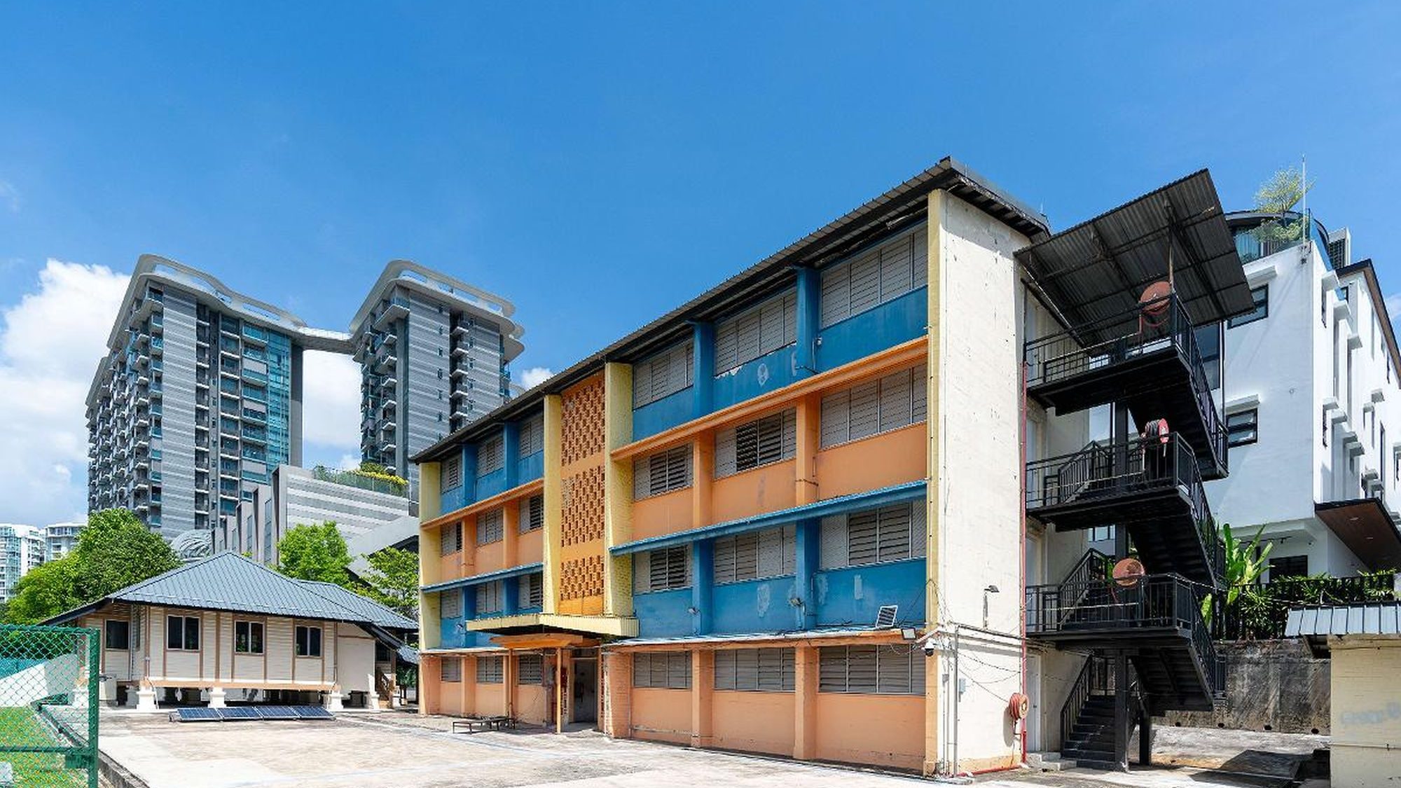 Three-storey older school building painted blue and orange with fire escape stairs and nearby small wooden house