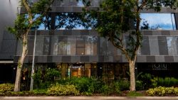 Commercial building facade with reflective windows, greenery in front, and signs for Caralell and Namu at street level