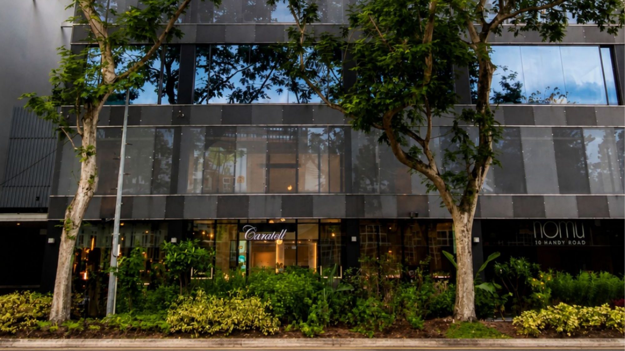 Commercial building facade with reflective windows, greenery in front, and signs for Caralell and Namu at street level