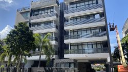 Mid-rise residential apartment blocks with balconies, surrounded by palm trees and a worker on a lift cleaning the building facade