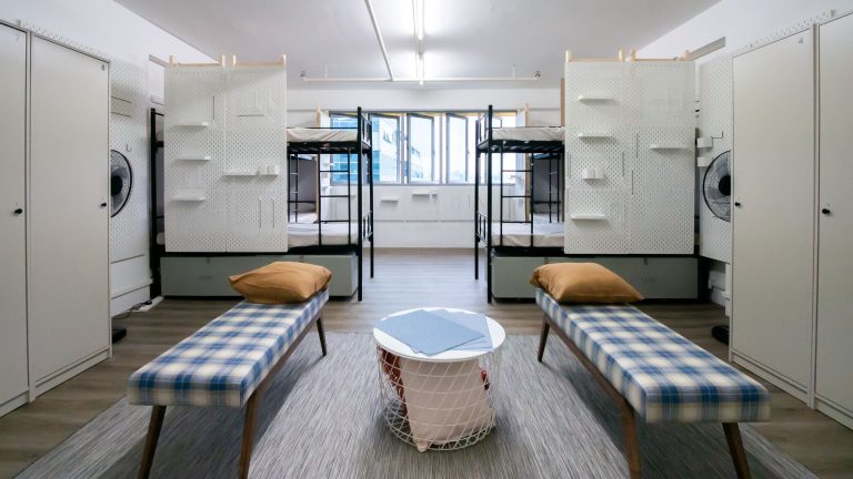 Room with two sets of metal bunk beds, plaid benches with brown pillows and lockers along the walls