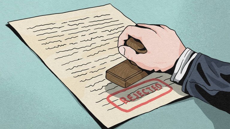 Hand holding a wooden stamp marking the word "REJECTED" in red on a document with text