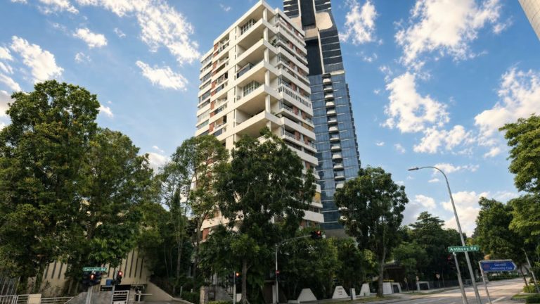 White residential apartment block with balconies beside a tall blue glass building and surrounding trees