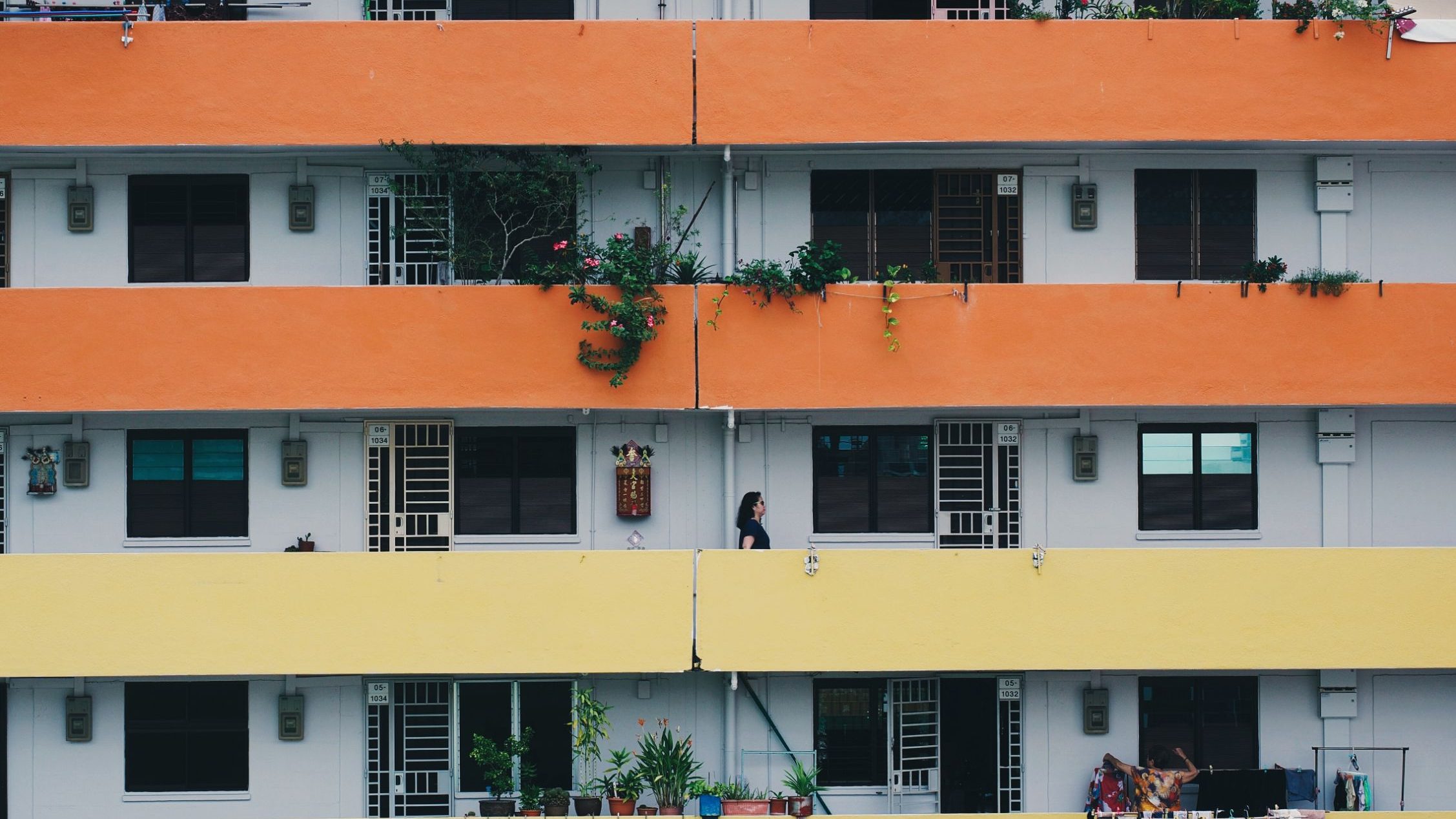 Housing block with orange and yellow corridors, plants on balconies, and people walking and drying clothes