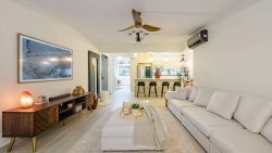Living room with a long white sofa, wooden TV console, ceiling fan, and open kitchen with bar stools