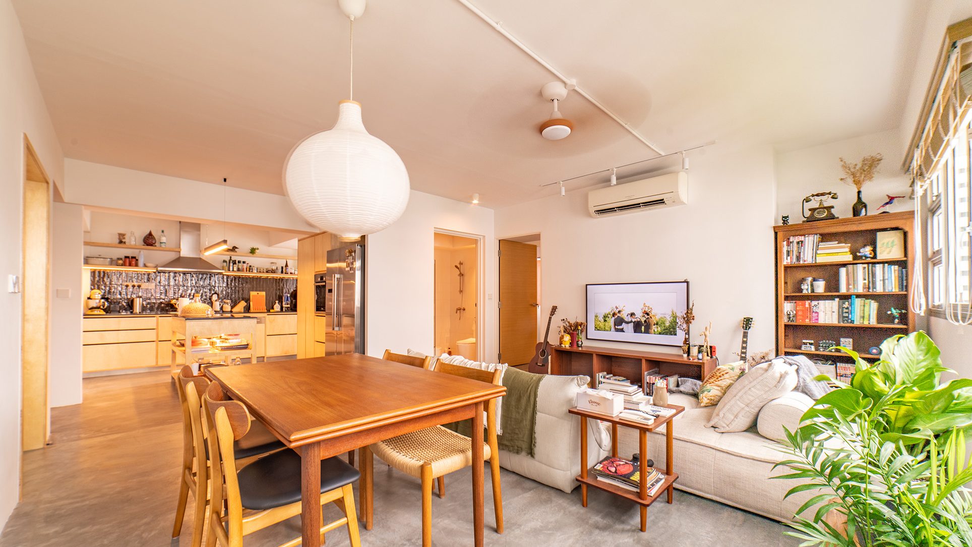 Dining and living area with wooden table and chairs, cream sofa, TV console, bookshelf, and open kitchen background