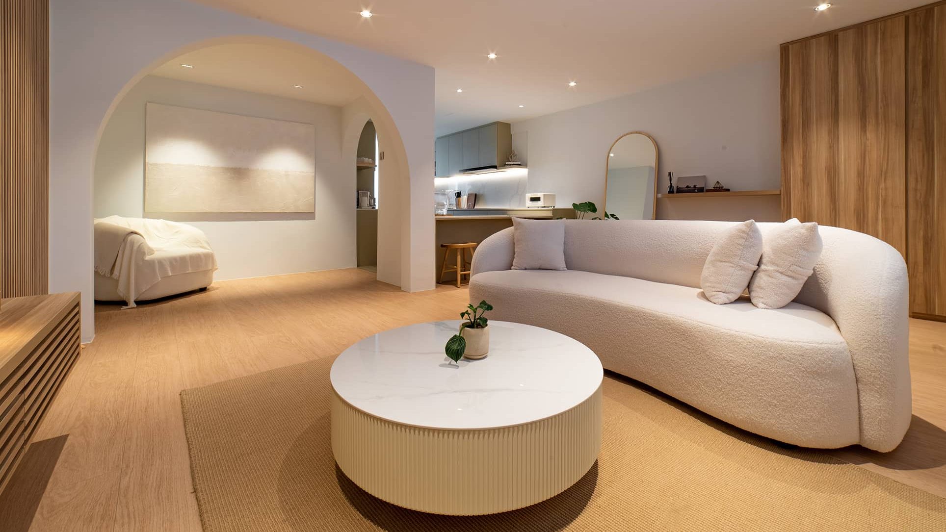 Living room with curved white sofa, round marble coffee table, wood panel wall and kitchen in background