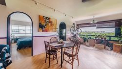 Dining area with wooden table and chairs, plants by large window, and bedroom visible through arched doorway