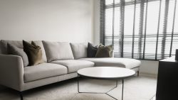 Living room with light grey L-shaped sofa, dark cushions, round white table and window blinds