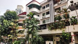 Four-storey residential block with curved balconies, plants in front, and high-rise buildings in distance