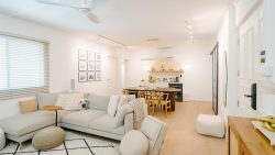 Living room with beige sectional sofa and dog face cushion, beside a dining table with wooden chairs and shelving unit