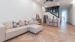 Living room with beige sofa set, light wood herringbone flooring, kitchen island with chairs, and ceiling fan