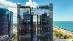 Tall glass-clad office towers with rooftop greenery near a coastal road and blue sea under a partly cloudy sky