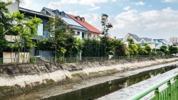 Row of terrace houses with tiled roofs behind trees along a concrete drainage canal with green railing