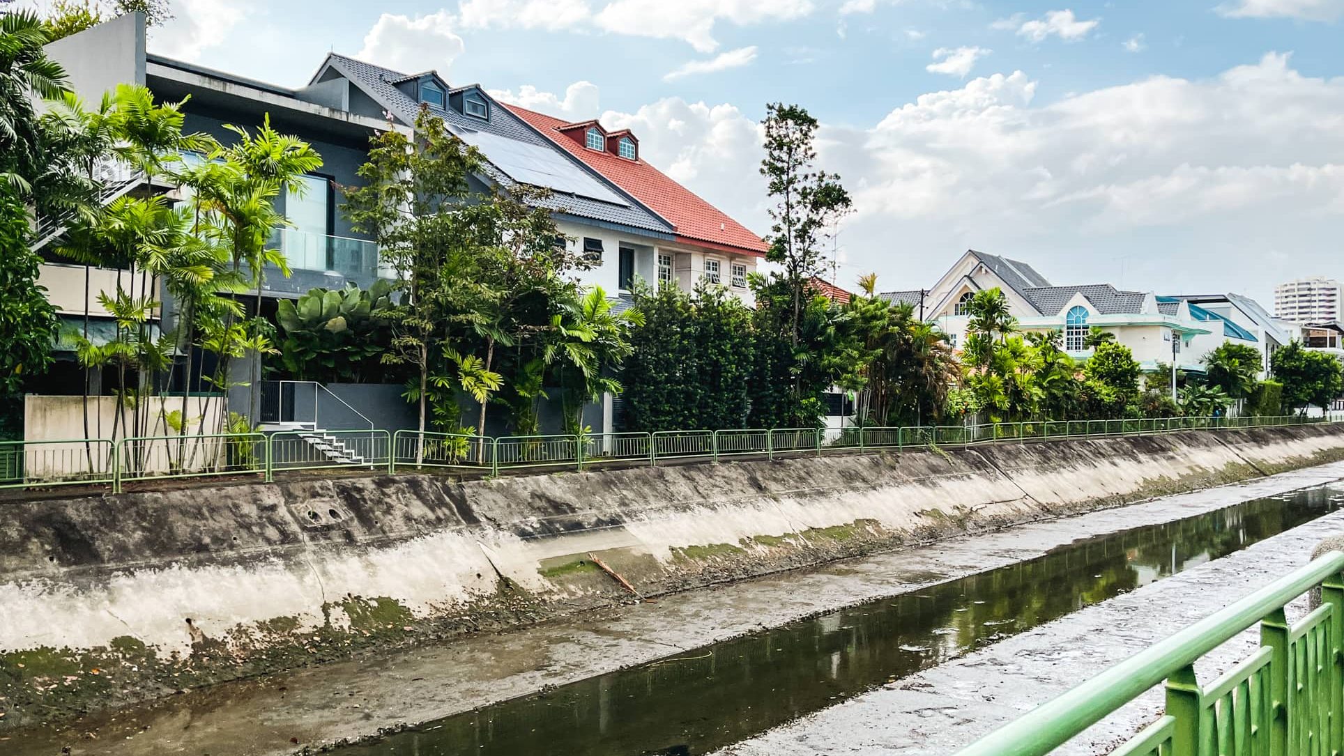 Row of terrace houses with tiled roofs behind trees along a concrete drainage canal with green railing