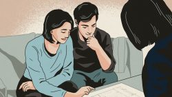 Couple seated on sofa reviewing house or apartment floor plan with a consultant or agent nearby
