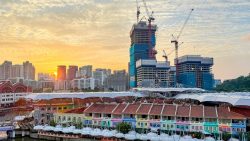 Low-rise shophouses with colourful façades by a river, high-rise buildings and construction cranes at sunset