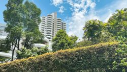 White residential tower with curved balconies behind green trees and hedges under a blue sky with clouds