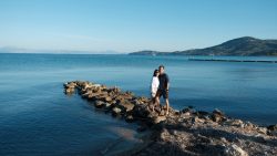 Couple standing on rocky shoreline with calm blue sea and mountain in the background
