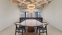 Meeting room with a long wooden table, black chairs, hanging lights, and cityscape windows at the end