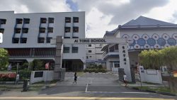 Entrance gate and buildings of Ai Tong School with people walking nearby and cloudy sky above
