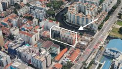 Cluster of mid-rise residential blocks with red tiled roofs beside a river and main road with arrow and outline highlight