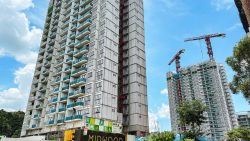 Two high-rise residential blocks under construction with cranes and greenery under a blue sky