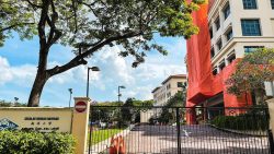Main school building partially covered in red protective netting behind gate with Sekolah Rendah Nanyang signboard