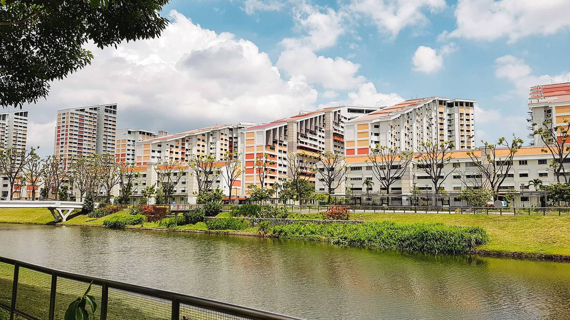 Low-rise and high-rise housing blocks near a river with a footbridge and trees on a sunny day