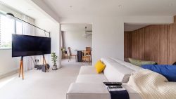 Living room with cream sofa, colourful cushions, TV on wooden legs, and dining area with wooden chairs in background