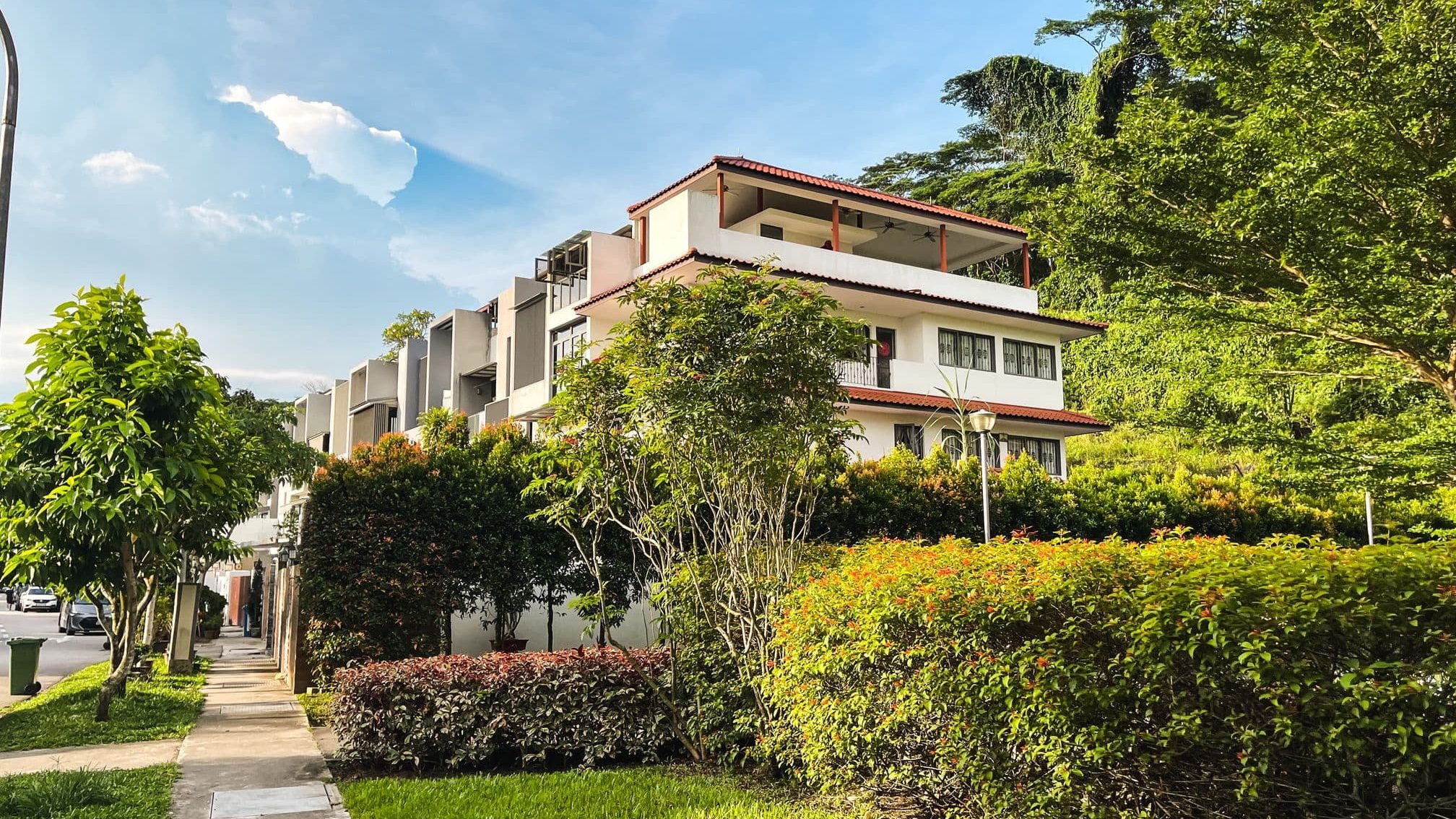 Three-storey terrace houses with tiled roofs along a tree-lined sidewalk and lush greenery