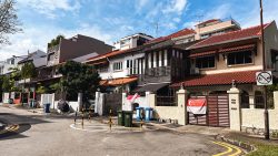 Two-storey terrace houses with red tiled roofs along a curved street displaying Singapore flags on gates