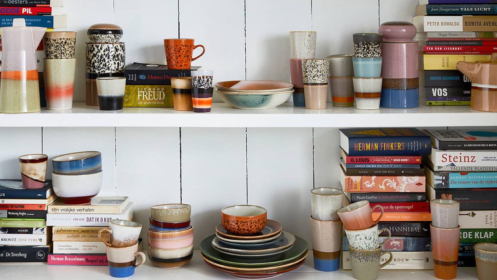 Shelves with colourful ceramic mugs, bowls, plates and stacks of books against a white wooden wall