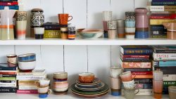 Shelves with colourful ceramic mugs, bowls, plates and stacks of books against a white wooden wall